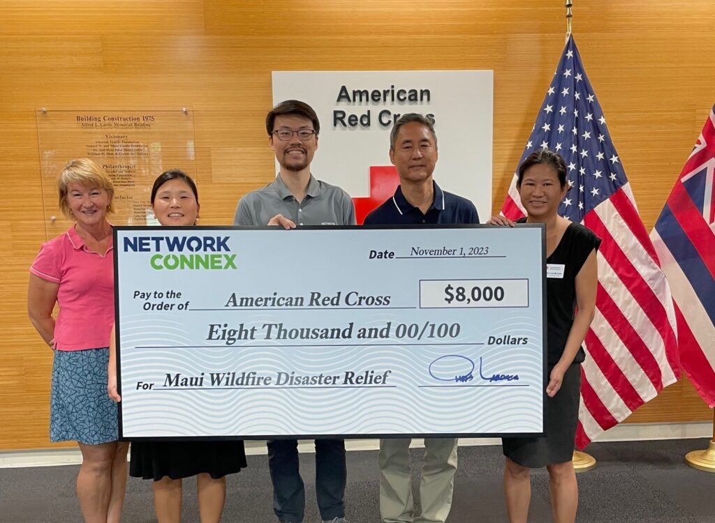 A group of five people standing in front of US and Hawai'i flags holds a large check from Network Connex written to the American Red Cross in the amount of $8,000